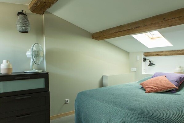 Une chambre avec un puits de lumière, un lit recouvert d’une courtepointe turquoise, une commode avec des tiroirs, une lampe fixée au mur et un luminaire suspendu.