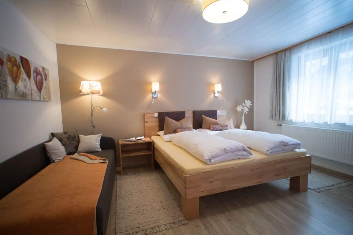 A bedroom with a bed, a sofa, a nightstand, wall-mounted lamps, and a window with curtains.