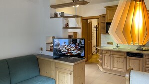 A living room with a television displaying a family meal, a sofa, wooden cabinets, and a kitchen area with a unique pendant light. - Dormio Gasthof Höllwirt (Obertraun)