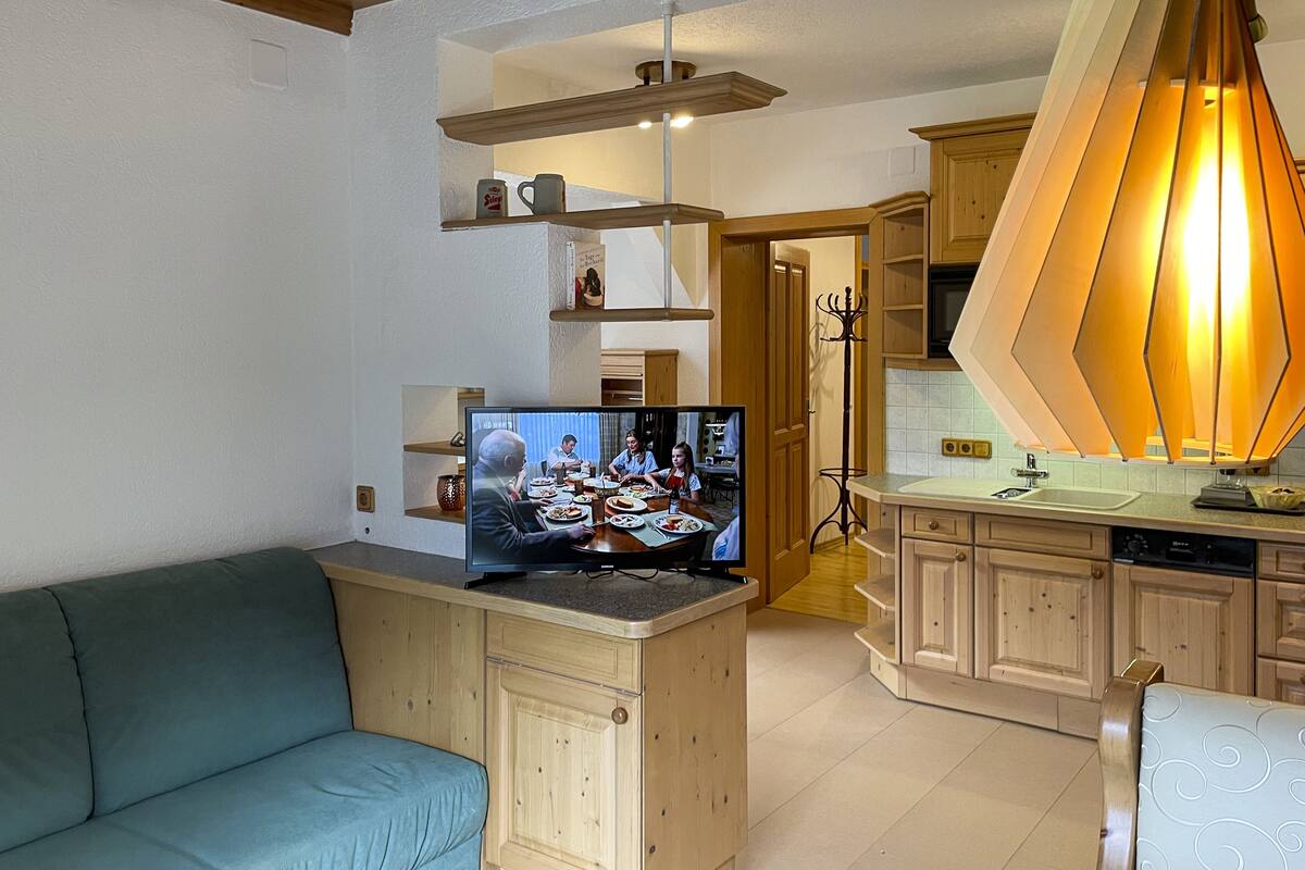 A living room with a television displaying a family meal, a sofa, wooden cabinets, and a kitchen area with a unique pendant light.