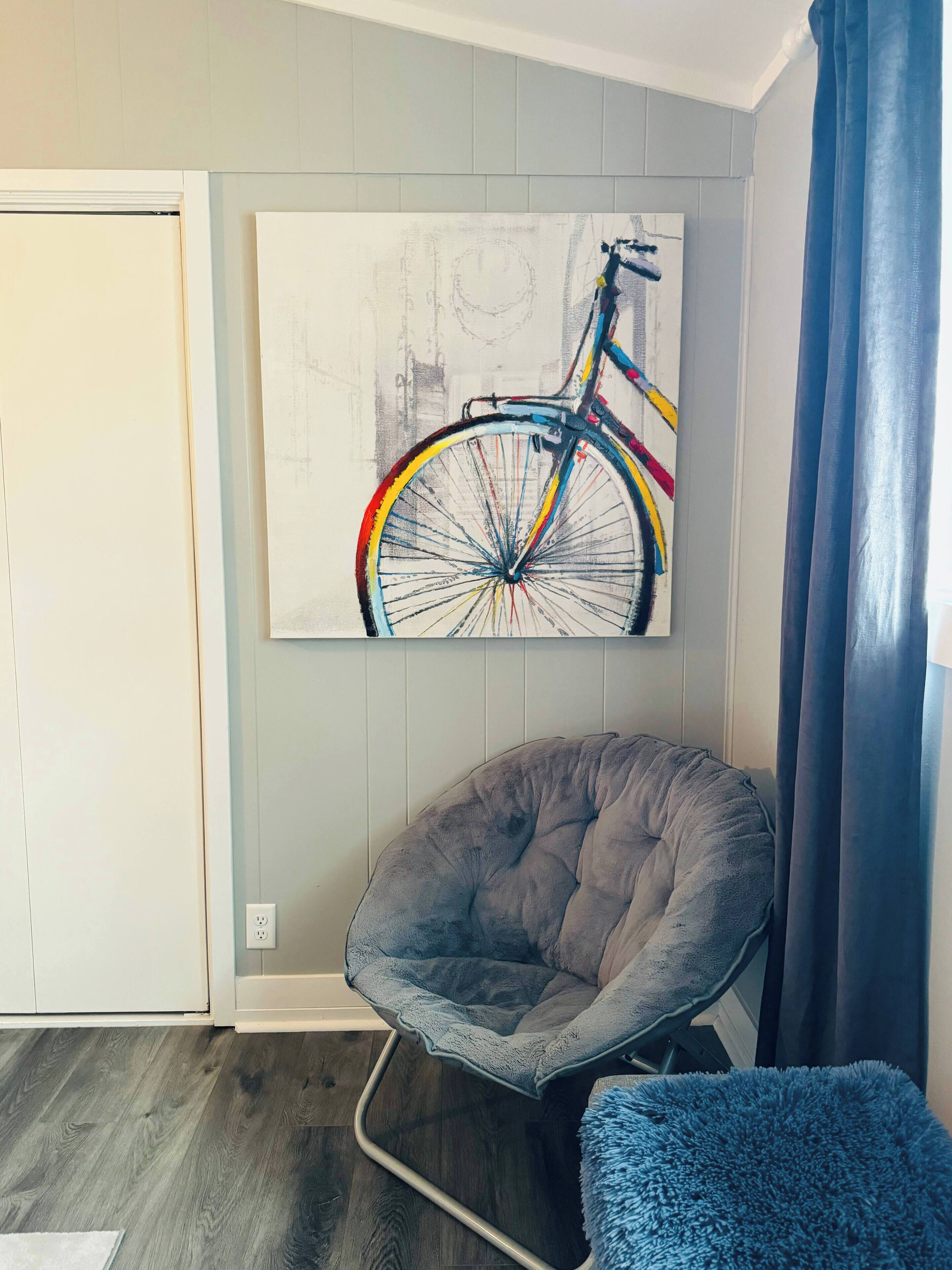 Une pièce comprenant un fauteuil à revêtement gris, un pouf bleu et un tableau représentant un vélo, accroché au mur.