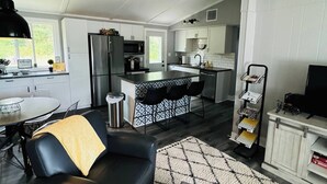 Une cuisine moderne avec des armoires blanches, un réfrigérateur noir et un four à micro-ondes. On y trouve un coin repas avec une table et des chaises, ainsi qu’un canapé en cuir noir avec un plaid. Un pouf gris se trouve au centre de la pièce.