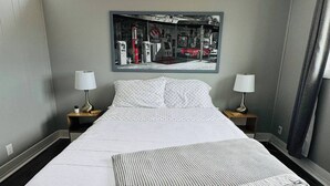 Une chambre d’hôtel avec un grand lit, deux tables de chevet avec des lampes, une fenêtre avec des stores et une œuvre d’art fixée au mur représentant une scène de station-service.