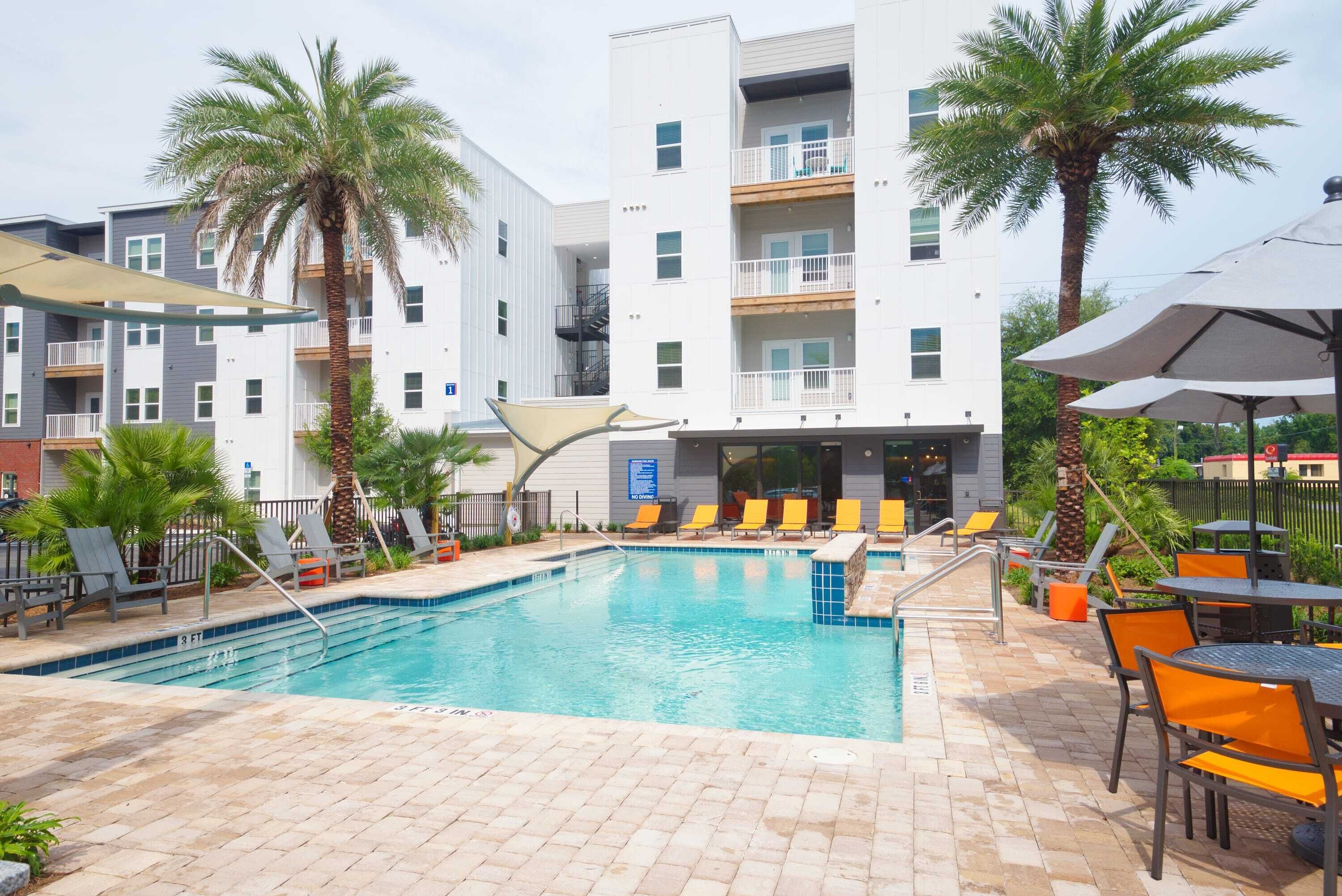 A swimming pool with lounge chairs and tables, surrounded by palm trees and a multi-story building with balconies.
