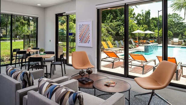 A modern indoor space with a pool view, featuring a dining area, lounge chairs, and a large window. - Walk to UF | Stylish 2BR | Ideal for Hospital & Gator Visitors (Gainesville)