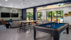 Une table de billard dans une pièce moderne, avec un canapé et des fauteuils, de grandes fenêtres et un plafond doté d’un éclairage encastré.