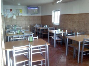 Classroom-style dining area with tables and chairs, a mounted TV, and brick walls.