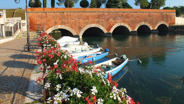 Un canal bordé de bateaux amarrés, un pont en briques et une rangée de lampadaires.