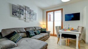 Un salon avec un canapé gris, une table à manger avec des chaises, un téléviseur fixé au mur et une porte donnant sur l’extérieur.
