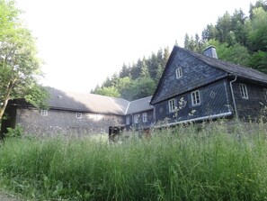 Une maison traditionnelle en bois, dotée d’un toit à forte pente, entourée d’une végétation luxuriante et d’arbres.
