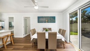 En matplats med ett vitt bord dukat för sex personer, omgiven av beige stolar, en takfläkt och ett stort fönster med utsikt över grönskan.