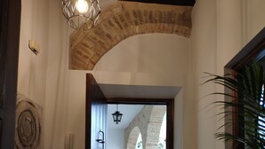 Ein Korridor mit einem steinernen Bogen, der zu einem Außenbereich mit einer Backsteinmauer und einem Blumentopf führt.
