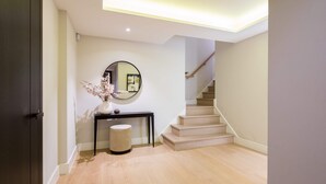 A modern hallway with a wooden floor, a round mirror on a console table, a small round stool, and a staircase with beige steps.