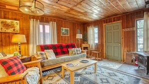 A cozy cabin interior with a wooden ceiling, a sofa with a red and black checkered blanket, a coffee table, a fireplace, and a door. - Rustic Riverside Cabin with Patio — Family Escape (Lyons)