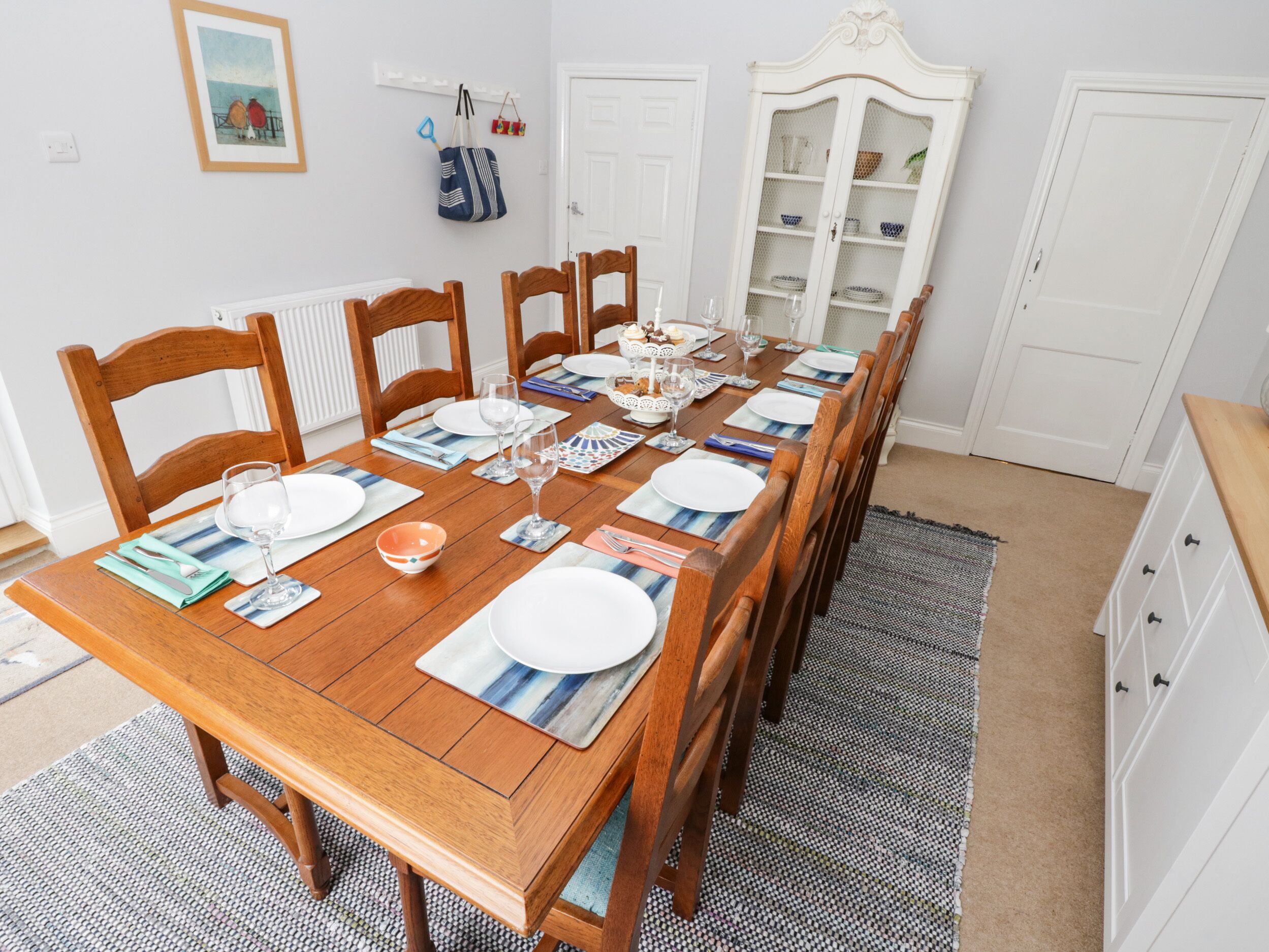 A dining area with a wooden table set for six, surrounded by wooden chairs. There is a white cabinet with glass doors in the background.