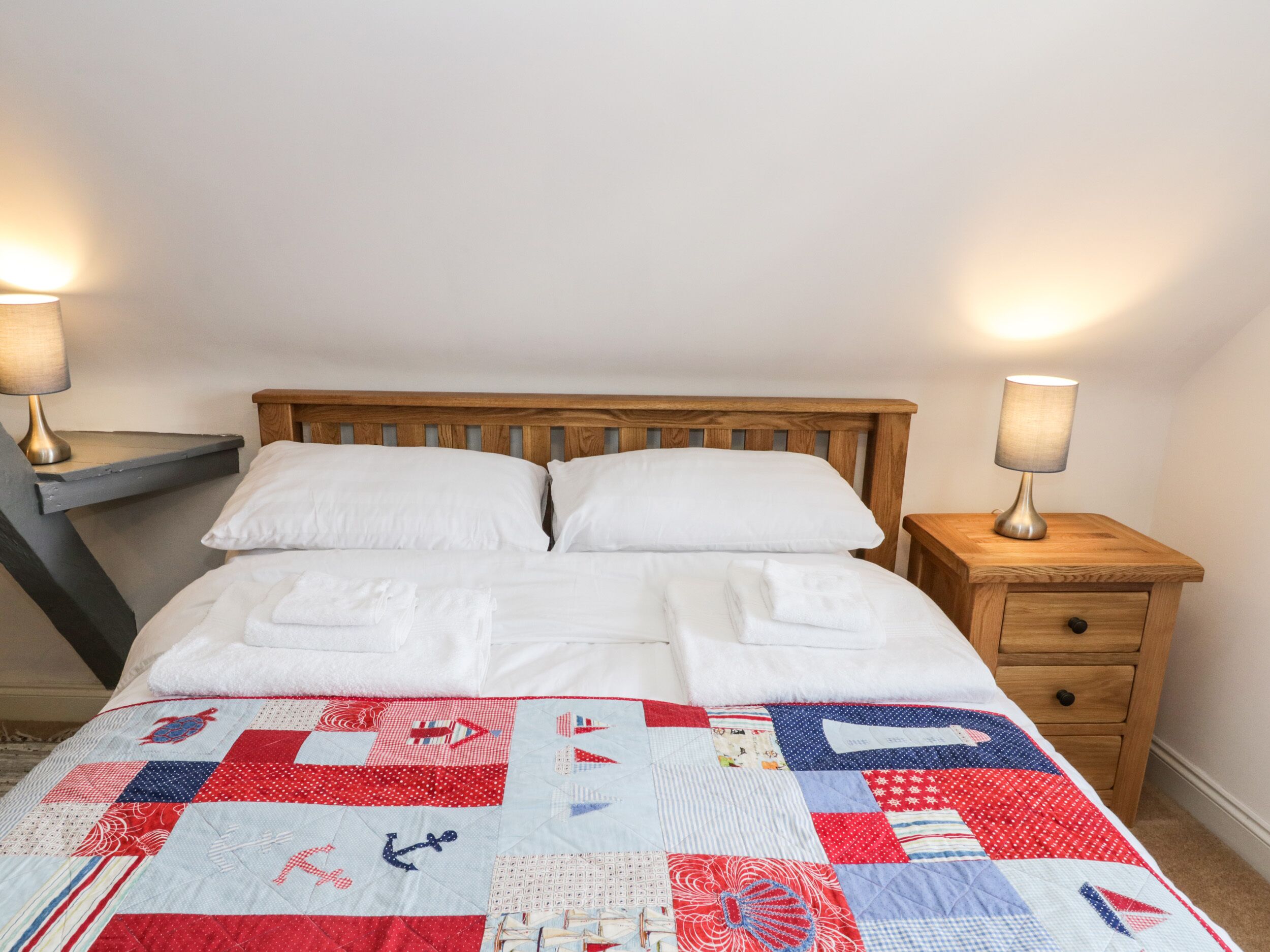 A neatly made bed with white linens and a red, white, and blue quilt. There are two wooden nightstands with lamps on either side of the bed.