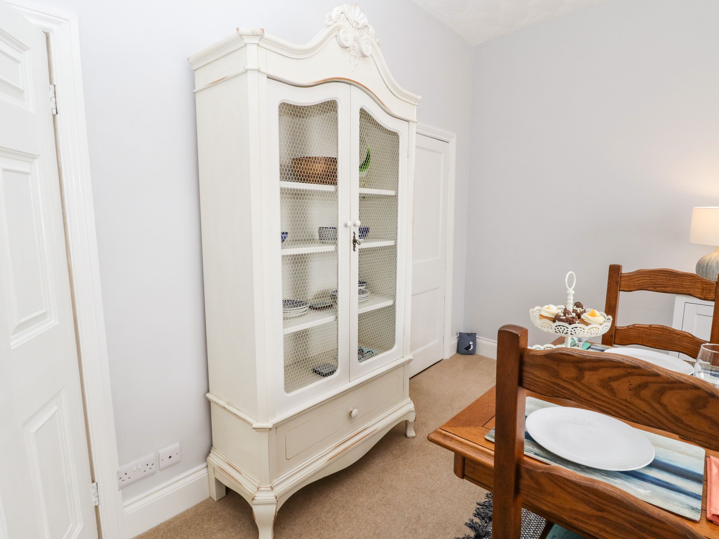A room with a white armoire, a wooden dining table with a plate, and a lamp.