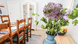 Uma área de refeições com mesa de madeira, cadeiras, um vaso com flores e uma cesta de frutas.