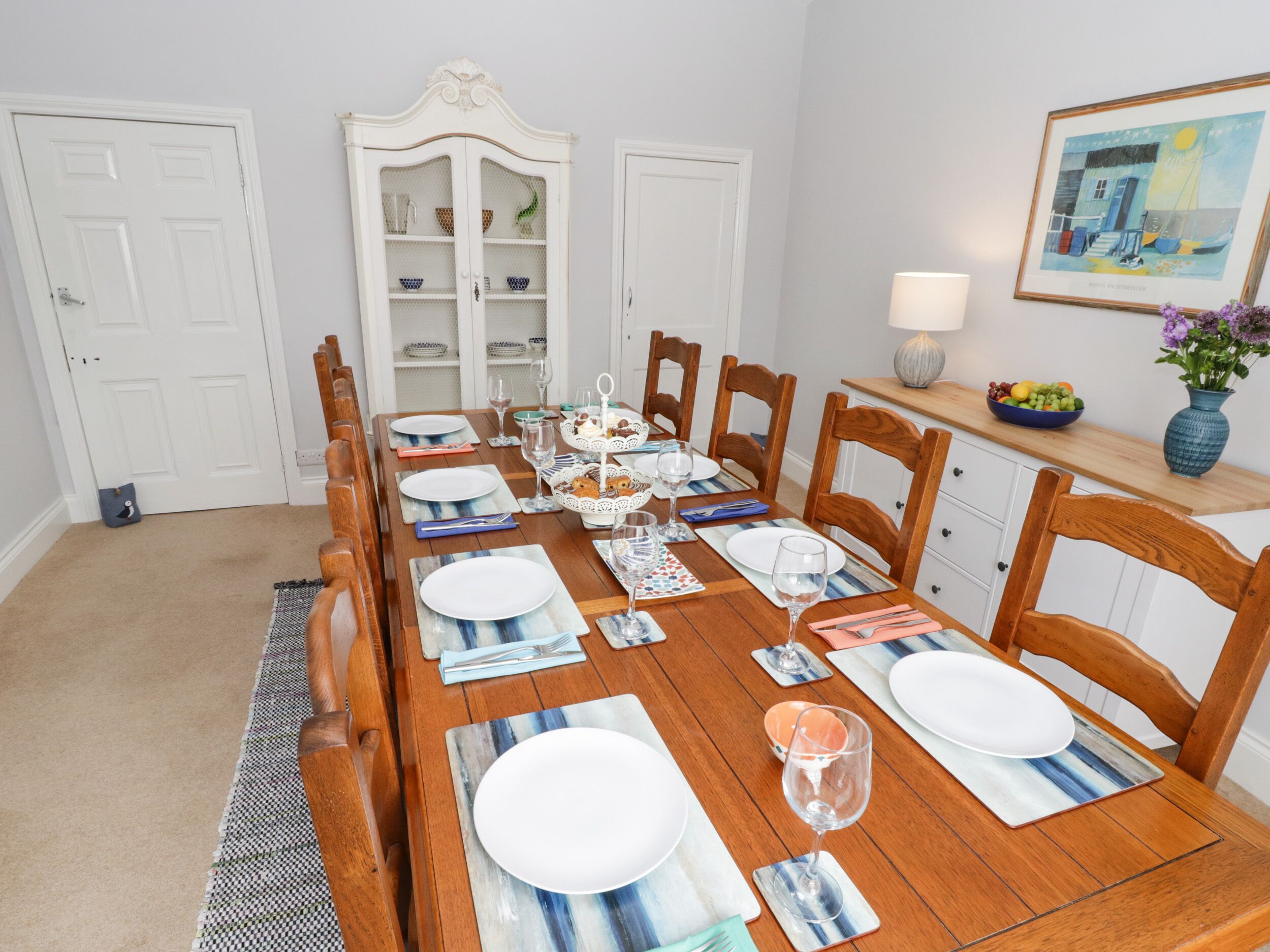 A dining room with a wooden table set for six, surrounded by chairs. There is a white cabinet with glass doors against the wall, and a framed picture above the table.