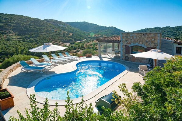 Relax by the oval-shaped pool surrounded by lush green hills and shaded by a pergola.