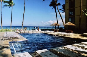 A pool area with lounge chairs and umbrellas, surrounded by palm trees and overlooking the ocean.