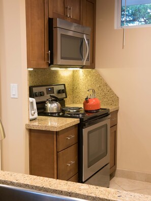 A kitchen with wooden cabinets, a stainless steel microwave, a gas stove, and a red kettle. - Oceanfront Mahana Condo  1BR/1BA Sweeping Views Ac, Pool (Lahaina)