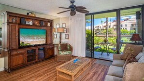 En stue med stort fladskærms-tv, tv-møbel i træ, fletsofa og en balkon med udsigt til bygninger og grønt.