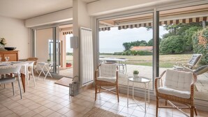 Et rummeligt opholdsområde indendørs med en stor skydedør, der fører ud til en udendørs terrasse. Her er fletstole, et spisebord og et lille hvidt bord.