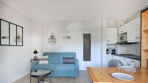A modern living room with a blue sofa, a round coffee table, and a dining table set for two. A kitchen is visible in the background with white cabinets and a microwave. - Apartment for 2 adults/2 kids, balcony (La Baule-Escoublac)