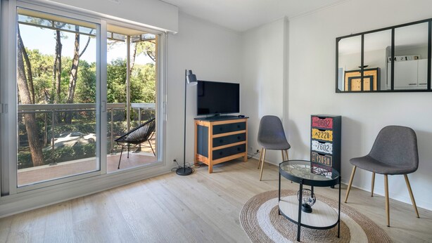 A modern living room with a large sliding glass door, a flat-screen TV on a wooden stand, two grey chairs, a small round table, and a decorative mirror on the wall. - Apartment for 2 adults/2 kids, balcony (La Baule-Escoublac)