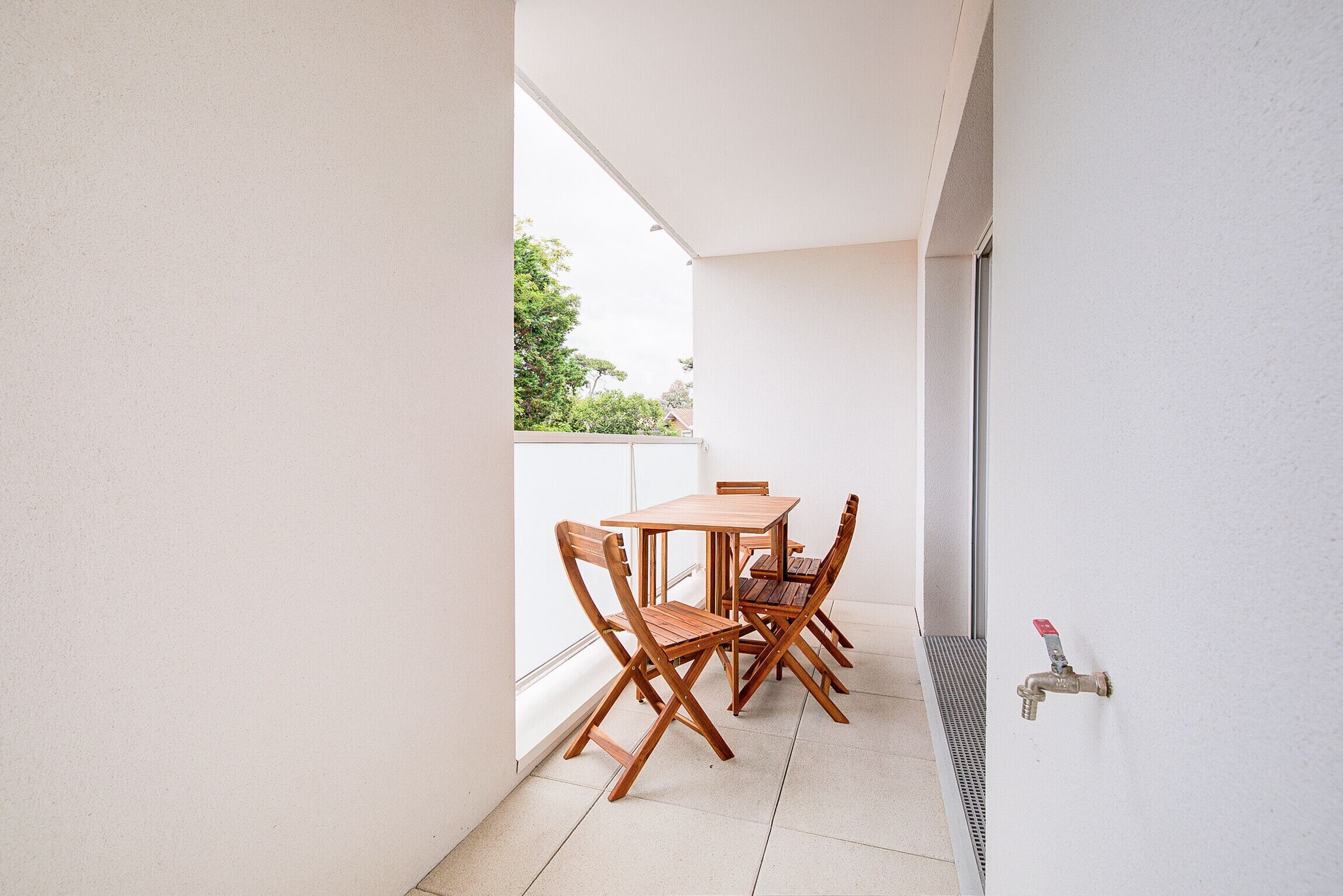 Un petit balcon avec une table et des chaises en bois, une fenêtre et un robinet.