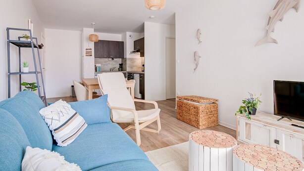 A modern living room with a blue sofa, a wooden chair, a TV, and a kitchen in the background. - Relax in this beautiful flat in Lacanau (Lacanau)