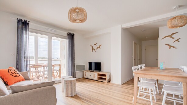 A modern living room with a sofa, a dining table with chairs, a TV, and a view of the outdoors.