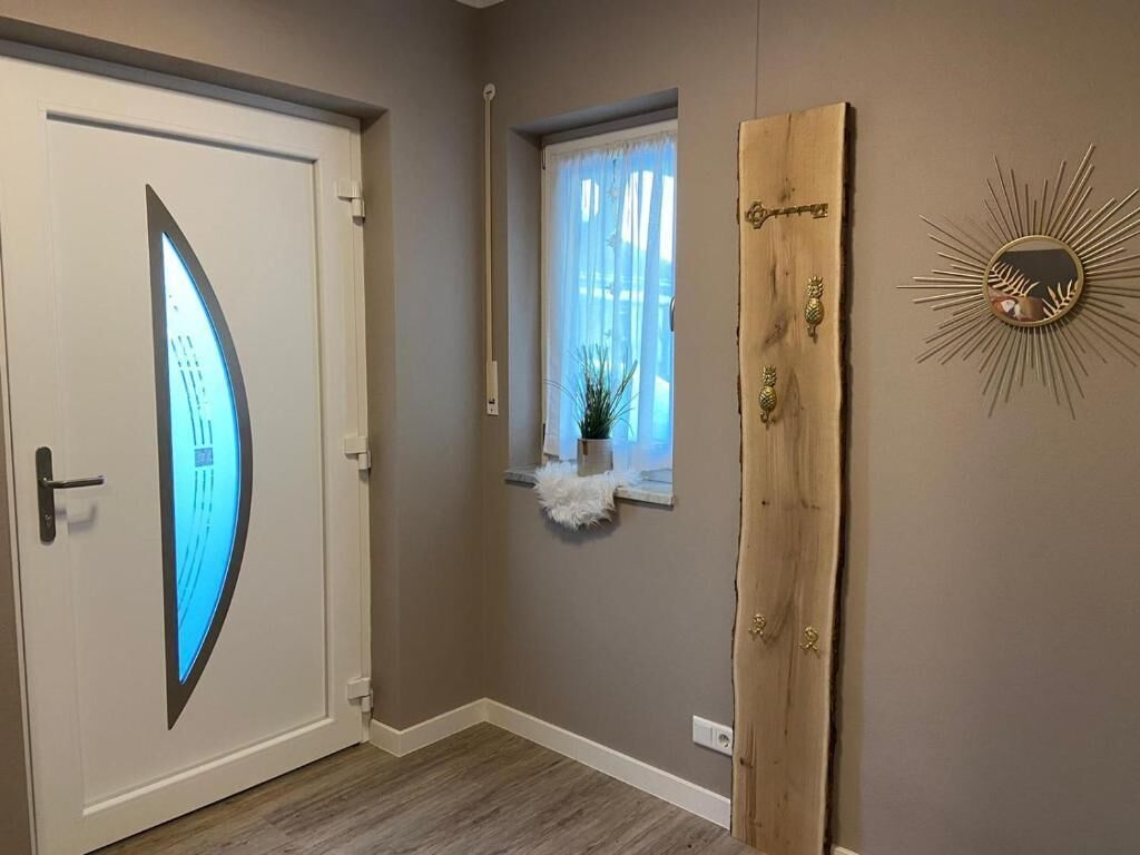 A hallway with a white door featuring a blue light, a small window with a white curtain, and a wooden panel with a sunburst mirror on the wall.