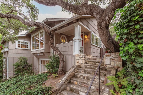 A two-story house with a stone staircase leading to a porch, surrounded by greenery and trees.
