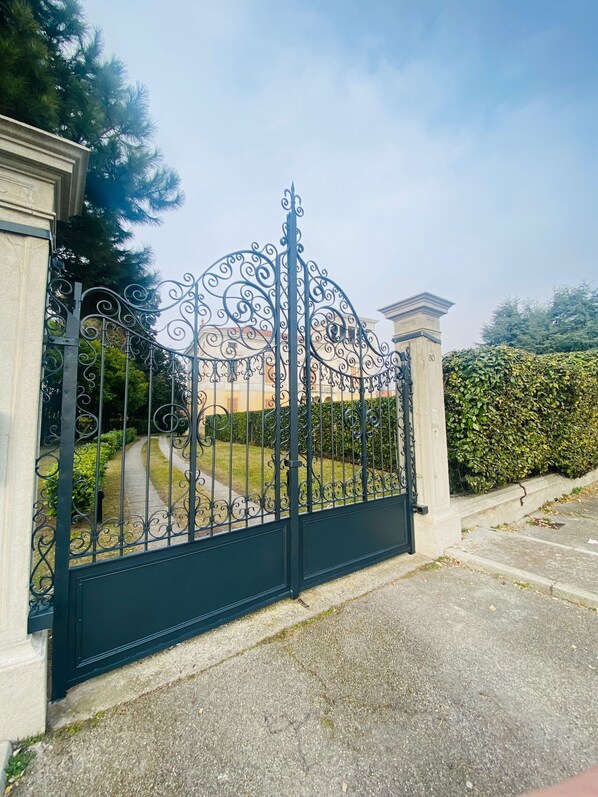 A black wrought iron gate with intricate designs, leading to a building with classical architecture and a well-maintained hedge.