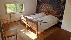 Une chambre à coucher avec deux lits en bois, une fenêtre donnant sur des palmiers, un radiateur et un mur orné d’un motif de feuilles.