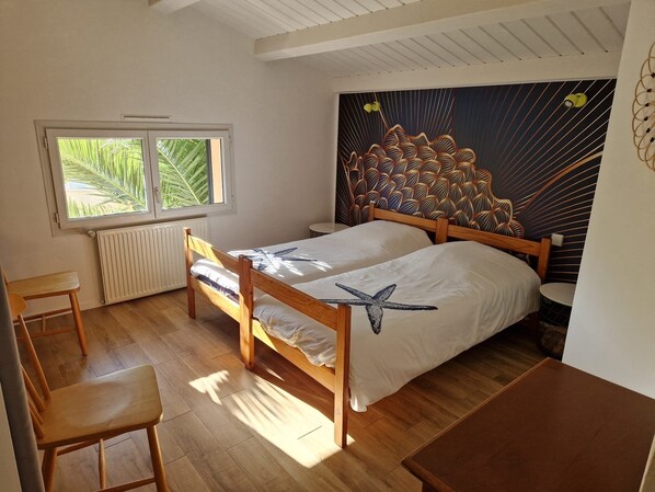 A bedroom with two wooden beds, a window with a view of palm trees, a radiator, and a wall with a leaf pattern.
