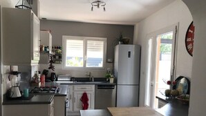 Une cuisine dotée d’un îlot central, de meubles blancs, d’un réfrigérateur et d’une fenêtre avec des stores.