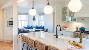 A modern kitchen with white cabinetry, a marble island, wicker bar stools, and pendant lighting. - Luxury Nantucket Estate | Heated Pool & Hot Tub (Nantucket)