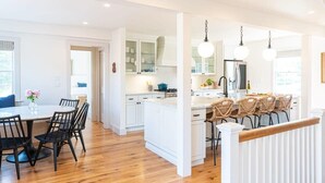 A modern kitchen with white cabinets, a central island, and a dining area with a round table and chairs. - Luxury Nantucket Estate | Heated Pool & Hot Tub (Nantucket)