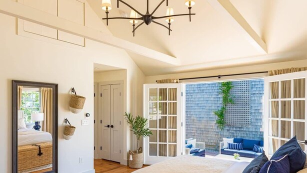 A bedroom with a large bed, a chandelier, a view of the outdoors, and a sliding door leading to a balcony. - Luxury Nantucket Estate | Heated Pool & Hot Tub (Nantucket)