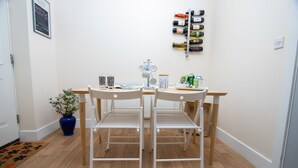 Une pièce comprenant deux chaises en métal blanc et une table en bois, un vase bleu avec des fleurs blanches, un porte-bouteilles fixé au mur et une porte avec un cadre blanc.