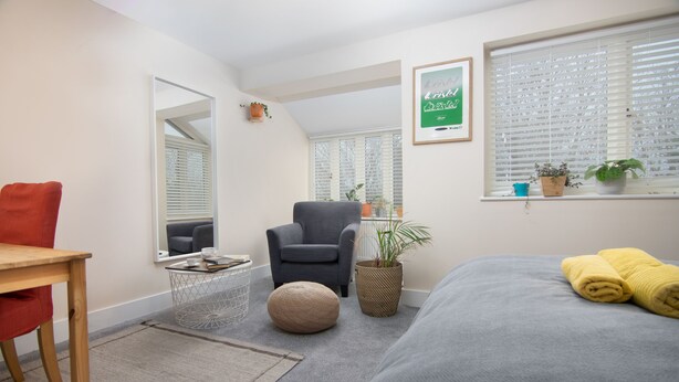 Une chambre d’hôtel moderne avec un lit gris, une chaise rouge, un fauteuil gris, un petit pouf rond, une table d’appoint avec une plante et une fenêtre avec des stores.