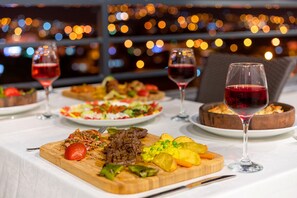 A dining table with various dishes and wine glasses, set against a backdrop of blurred city lights. - Artes Hotel (Defne)