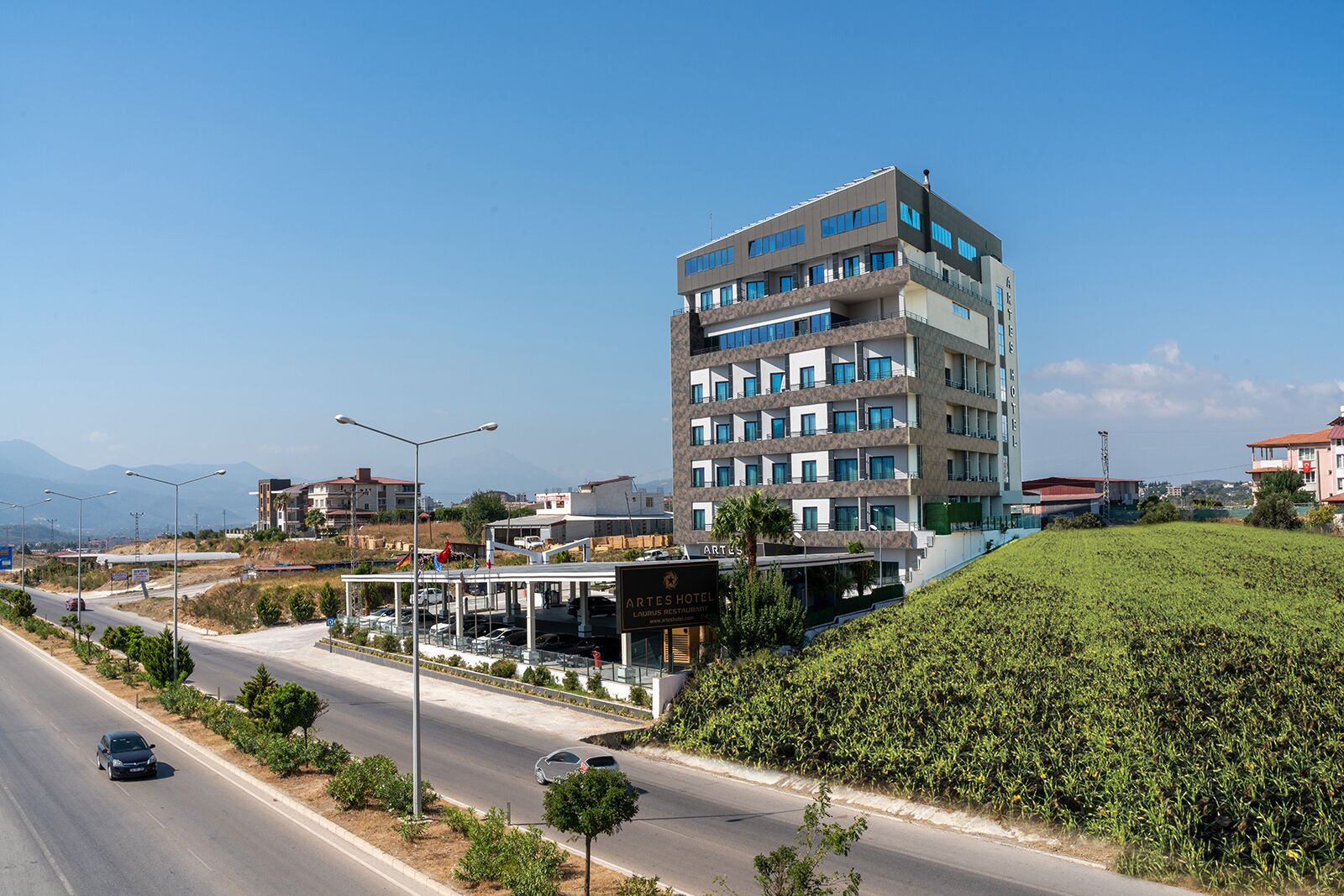 A multi-story hotel with a modern design, located adjacent to a road with a landscaped area and a view of distant mountains.