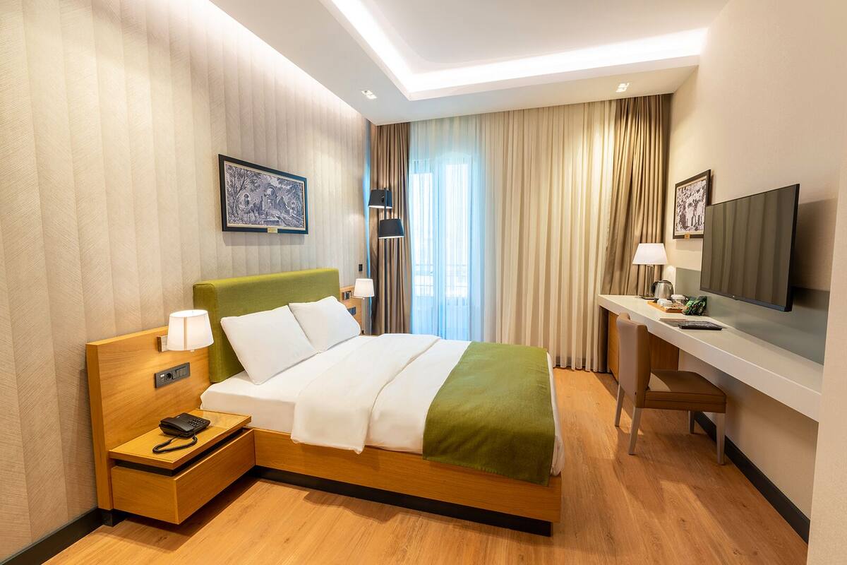 A hotel room with a bed, bedside table with a phone, a desk with a TV, and a window with curtains.