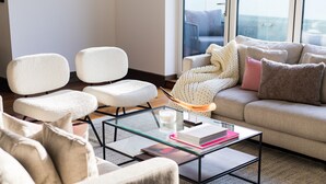 En moderne stue med et glasbord, to hvide lænestole, en beige sofa med puder og udsigt gennem et stort vindue.