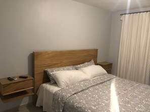 Ein Schlafzimmer mit einem Holz-Kopfteil, einem Bett mit gemusterten Betttextilien, einem Nachttisch mit Fernbedienung und einem Fenster mit Vorhängen.