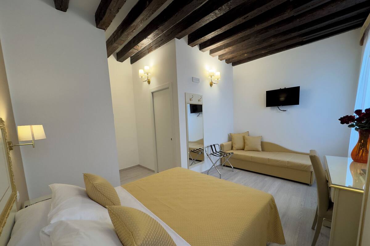 A hotel room with a bed, a sofa, a chair, a desk, a mirror, and a TV mounted on the wall.